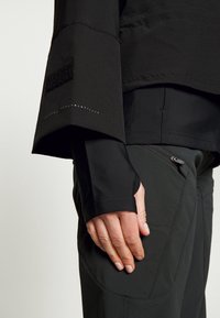 Top atlético negro con mangas anchas, que presenta acentos de velcro y detalles punteados. Combinado con pantalones gris oscuro, que exhiben un bolsillo con cremallera.