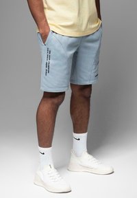 Shorts de sport bleu clair avec poches latérales, comportant des graphiques textuels noirs sur la jambe gauche et un tissu lisse et doux. Baskets blanches et chaussettes.