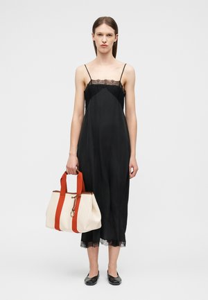 Žena v čiernych šatách s čipkovým lemom a čiernych balerínach, drží krémovú tašku na rameno so širokými oranžovými popruhmi a príveskom na kľúče.