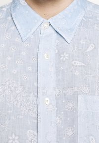 Jasnoniebieska, lniana koszula z krótkim rękawem z białym wzorem kwiatowym i paisley, z kołnierzykiem na guziki i kieszenią na piersi.