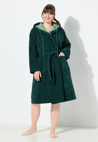 Accappatoio verde con cappuccio realizzato in tessuto morbido e testurizzato, dotato di una cintura da annodare e due tasche frontali; foderato con un materiale verde più chiaro.