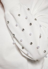 Felpa bianca con maniche decorate, caratterizzata da strass argentati e perle bianche, realizzata in tessuto testurizzato con una finitura morbida.
