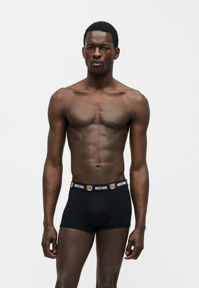 Homme torse nu, debout face à l'avant, portant un boxer noir avec le logo Moschino et des icônes d'ours en peluche sur la ceinture, sur un fond uni.