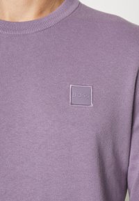 Pull violet en tricot avec un col rond et une étiquette carrée accentuée portant "BOSS" brodée au centre. Texture douce, coupe ajustée.