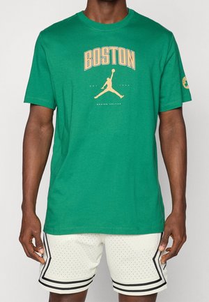 Mann trägt grünes Boston Celtics T-Shirt mit Basketballspieler-Logo und weißen perforierten Shorts mit schwarzem Rand.