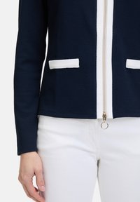 Veste zippée marine avec des accents blancs et deux poches frontales. Tissu texturé, design ajusté et détail de fermeture éclair doré. Portée sur un pantalon blanc.