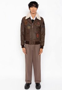 Rjava usnjena bomber jakna z ovratnikom iz ovčjega krzna, večimi našitki, sprednjim zadrgo in dvema velikima žepoma; kombinirana z bež hlačami.