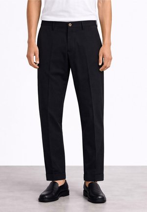 Uomo con pantaloni neri su misura con orli risvoltati e scarpe nere senza lacci, in piedi davanti a uno sfondo semplice.