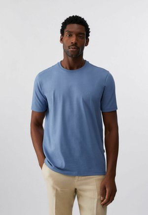 Mand med kort krøllet hår, iført en ensfarvet blå T-shirt med korte ærmer og beige bukser, stående foran en hvid baggrund.