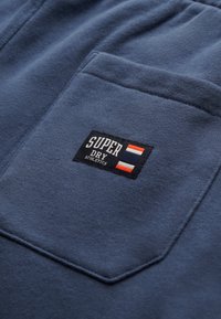 Mørkeblå sweatshirt med tekstureret stof og en lomme med lap. Har et sort mærke med "SUPERDRY" og tre striber i rød og hvid.