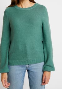 Grönstickad tröja med en texturerad design, rund hals, och långa ärmar, parat med ljusblå jeans.