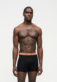 Μυώδης άνδρας που στέκεται μπροστά σε απλό φόντο, φορώντας μαύρα μποξεράκια Björn Borg με πορτοκαλί μέση.