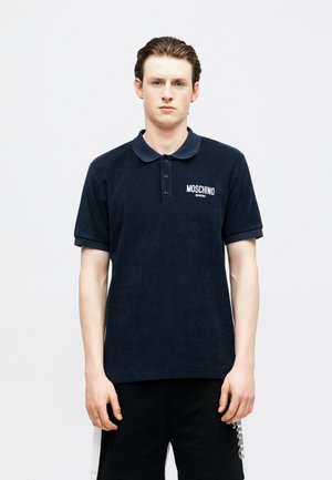 Mladý muž s vlasy uhladenými dozadu má na sebe námornícku modrú polokošeľu Moschino Swim a čierne šortky, stojí pred jednoduchým svetlým pozadím.