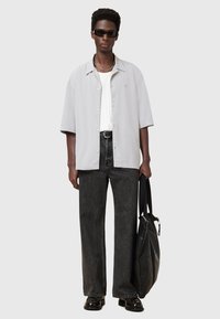 Camicia grigia chiaro a maniche corte, abbottonata, t-shirt bianca sotto, jeans neri a vita alta, scarpe nere, tiene una borsa nera, occhiali da sole.