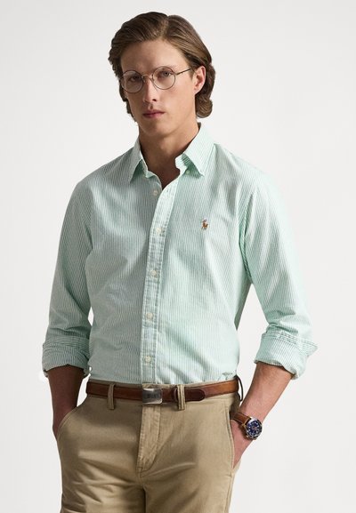 Camisa de rayas verde claro con cuello abotonado, mangas remangadas, pequeño logo bordado, combinada con pantalones chinos khaki y cinturón marrón.