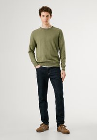Suéter verde oliva con un cuello redondo clásico, puños acanalados, combinado con jeans azul oscuro y zapatos marrones de cordones. Fondo liso.