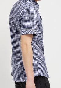 Koszula z krótkim rękawem w niebiesko-białą kratę, z kołnierzykiem na guziki i pojedynczą kieszenią na piersi. Materiał bawełniany.