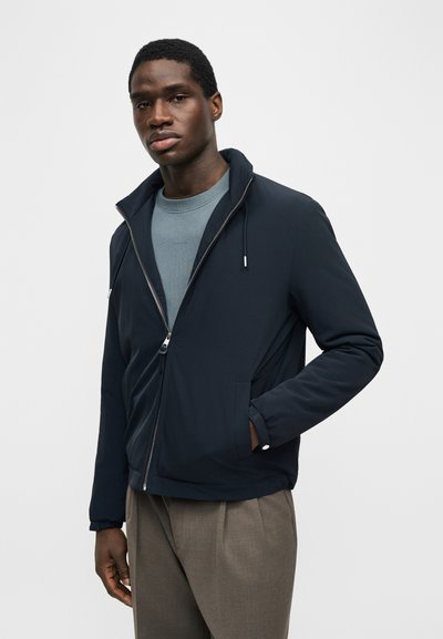 Άνδρας με μπλε μπουφάν με φερμουάρ πάνω από μπλε πουκάμισο και καφέ παντελόνι, στέκεται με το ένα χέρι στην τσέπη με φόντο απλή επιφάνεια.