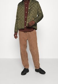 Grön vadderad jacka med stort diamantmönster, bruna kordbyxor och svarta sneakers, bärs över en vinröd knapp-up skjorta.