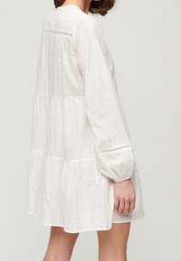 Kvinde iført en hvid, langærmet, lagdelt kjole med blonderdetaljer på ærmerne og ryggen, vist fra siden mod en ensfarvet baggrund.