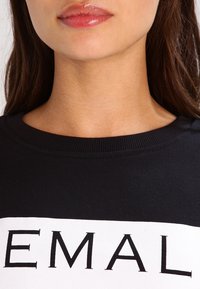 Svart sweatshirt med ett brett vitt band med svart text. Har ribbad ringning och mjuk tygstruktur.