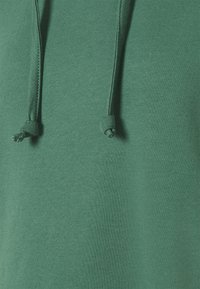 Detailní záběr na zelenou látku mikiny s kapucí, s dvěma šňůrkami volně visícími dolů, zobrazující texturu a detaily šití.