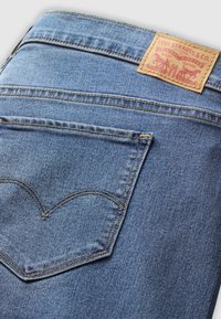 Denimjeans i mellomblått, med en signaturstikking på baklommen og en brun lærmerkelapp med rød tekst og grafikk.