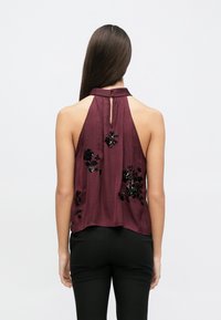 Μπουζάκι μπορντό με λαιμόκοψη halter και άνοιγμα στην πλάτη, διακοσμημένο με μαύρη κεντητή λουλουδένια παγιέτα. Λείο ύφασμα, άνετη εφαρμογή, σε συνδυασμό με μαύρο παντελόνι.