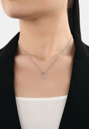 Silberne Halskette mit einem kleinen teal-farbenen Anhänger in Tropfenform und einem dekorativen Gliederdetail. Getragen zu einem schwarzen Blazer.