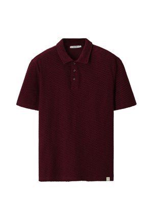 Polo bordeaux à manches courtes en tissu texturé, patte à trois boutons, et petite étiquette blanche au bas de l’ourlet.