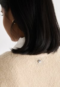 Beige knitted sweater with a textured fabric, featuring a small silver logo badge on the back near the neckline. White collared shirt underneath.