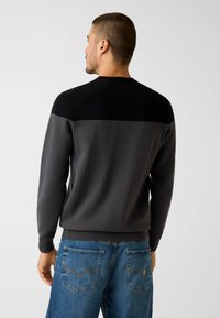 Grau-schwarz getönter Pullover mit Rundhalsausschnitt, langen Ärmeln und geripptem Saum. Kombiniert mit blauen Jeans, die eine Detailgestaltung an den Gesäßtaschen aufweisen.