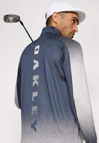 Navy og grå gradient golfjakke med høy krage, glidelås og "OAKLEY" trykt med store reflekterende bokstaver på ryggen.