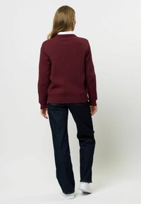 Pleteni maroon pulover s kabelskim vzorcem, rebrastimi manšetami in robom. V kombinaciji s temnimi kavbojkami in belimi supergami, prikazan od zadaj.
