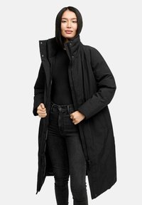 Kvinde med glat sort hår iført en lang sort polstret frakke over en sort top og sorte jeans, der holder frakken åben.