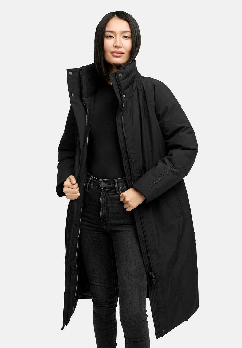 Kvinde med glat sort hår iført en lang sort polstret frakke over en sort top og sorte jeans, der holder frakken åben.