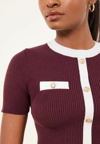 Punase tooniga soonikust top valgete detailidega, millel on ümmargune kaelus, lühikesed varrukad ja kuldsed nupud esiküljel ning rinnataskul.