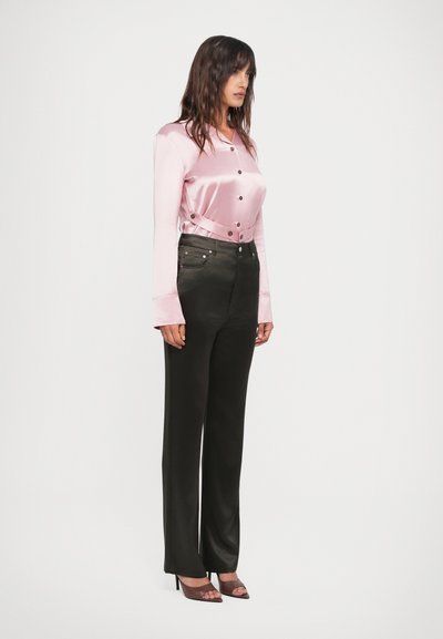Top lungo in satin rosa chiaro con maniche lunghe, bottoni e una cintura, abbinato a pantaloni neri a vita alta. Modella che indossa sandali trasparenti con tacco.