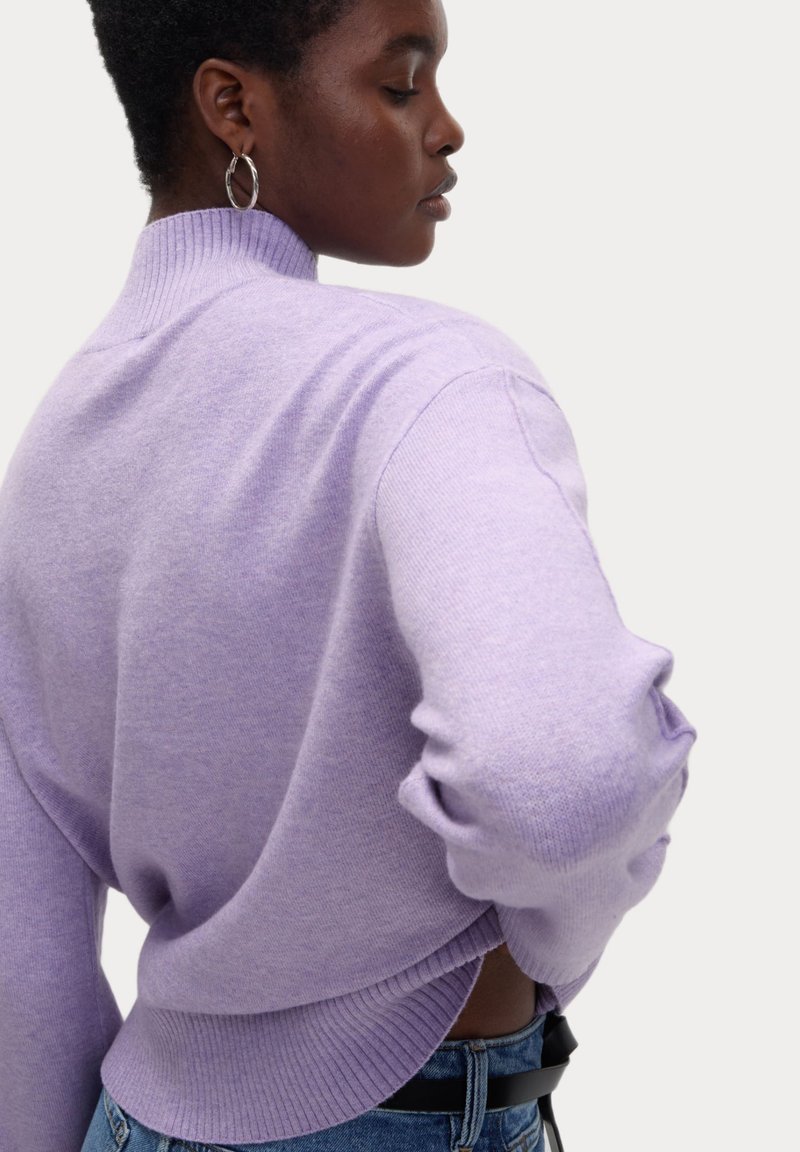 Lilafarbener Strickpullover mit hoch geripptem Kragen und lockerer Passform. Verfügt über gerippte Ärmelbündchen und einen gespaltenen Saum, kombiniert mit hochtaillierten Jeans.
