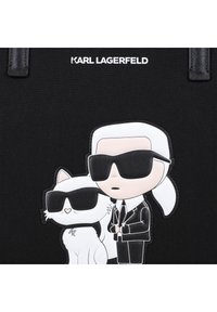 Μαύρη τσάντα tote με καρτουνίστικο σχέδιο ενός χαρακτήρα με γυαλιά ηλίου μαζί με μια άσπρη γάτα, και οι δύο σε παιχνιδιάρικη σχεδιαστική λεπτομέρεια.