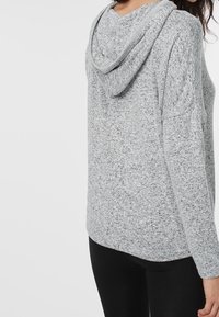 Sweat à capuche gris clair texturé avec capuche et épaules tombantes. Il présente un tissu tricoté doux et une coupe décontractée, associé à un legging noir.