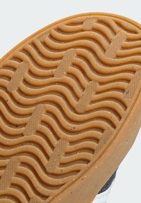 Hnědá gumová podrážka se zvlněným vzorem trakce a malými černými skvrnami. Boční pohled ukazuje krémové a námořnické části, které zdůrazňují výrazné designové prvky.