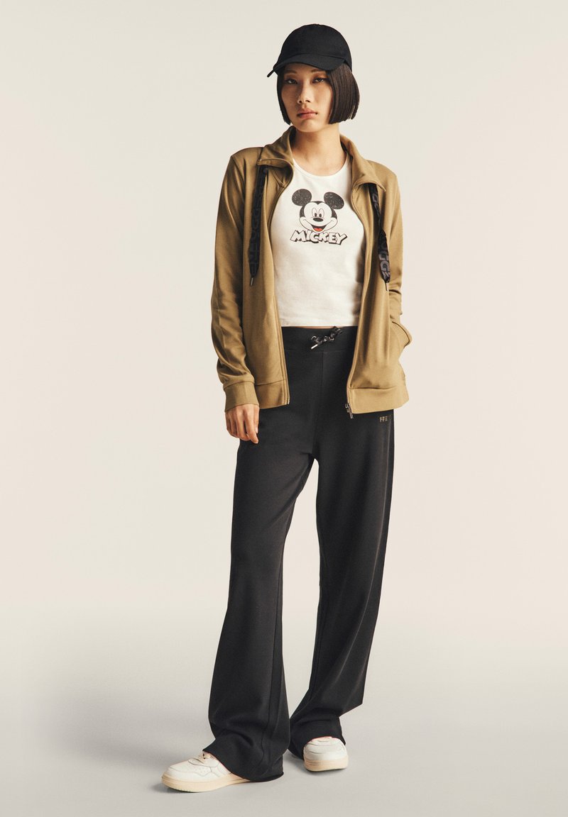 Chaqueta marrón con cremallera, camiseta blanca con gráfico de Mickey Mouse, pantalones negros de pierna ancha y zapatillas blancas. Una gorra negra completa el atuendo.