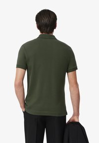 Polo shirt verde scuro a maniche corte, con colletto classico e tessuto testurizzato. Indossato con pantaloni neri, mentre tiene un giubbotto nero.