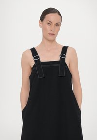 Rochie neagră fără mânici, confecționată dintr-un material neted, cu accesorii de culoare argintie pe bretelele reglabile și buzunare laterale.