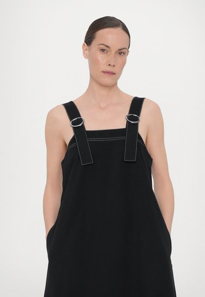 Crna haljina bez rukava izrađena od glatkog materijala, s metalnim detaljima srebrne boje na podesivim naramenicama i bočnim džepovima.