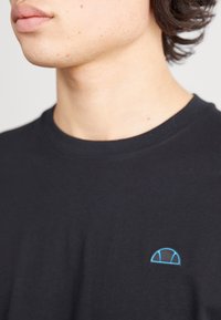 Černé bavlněné tričko s kulatým výstřihem. Na levé straně hrudi se nachází světle modré vyšívané logo v podobě oblouku. Hladká textura, snížený střih.