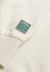 Tejido de sudadera color crema con una textura suave. Presenta un emblema acolchado verde con borde rosa, que muestra "SUPERDRY" y líneas cruzadas.