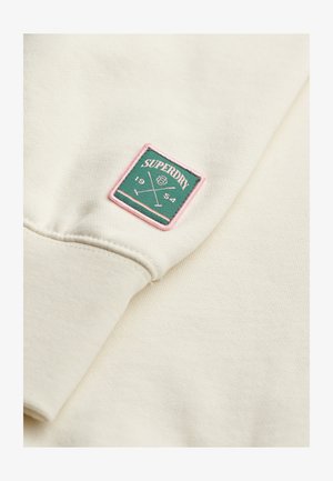 Cremfarbener Sweatstoff mit weicher Textur. Verfügt über ein grünes, gestepptes Emblem mit pinker Borte, auf dem "SUPERDRY" und gekreuzte Linien abgebildet sind.