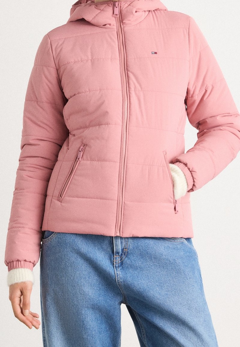 Veste matelassée rose avec capuche, poches zippées et poignets élastiques, portée avec un jean bleu clair, main partiellement dans la poche.